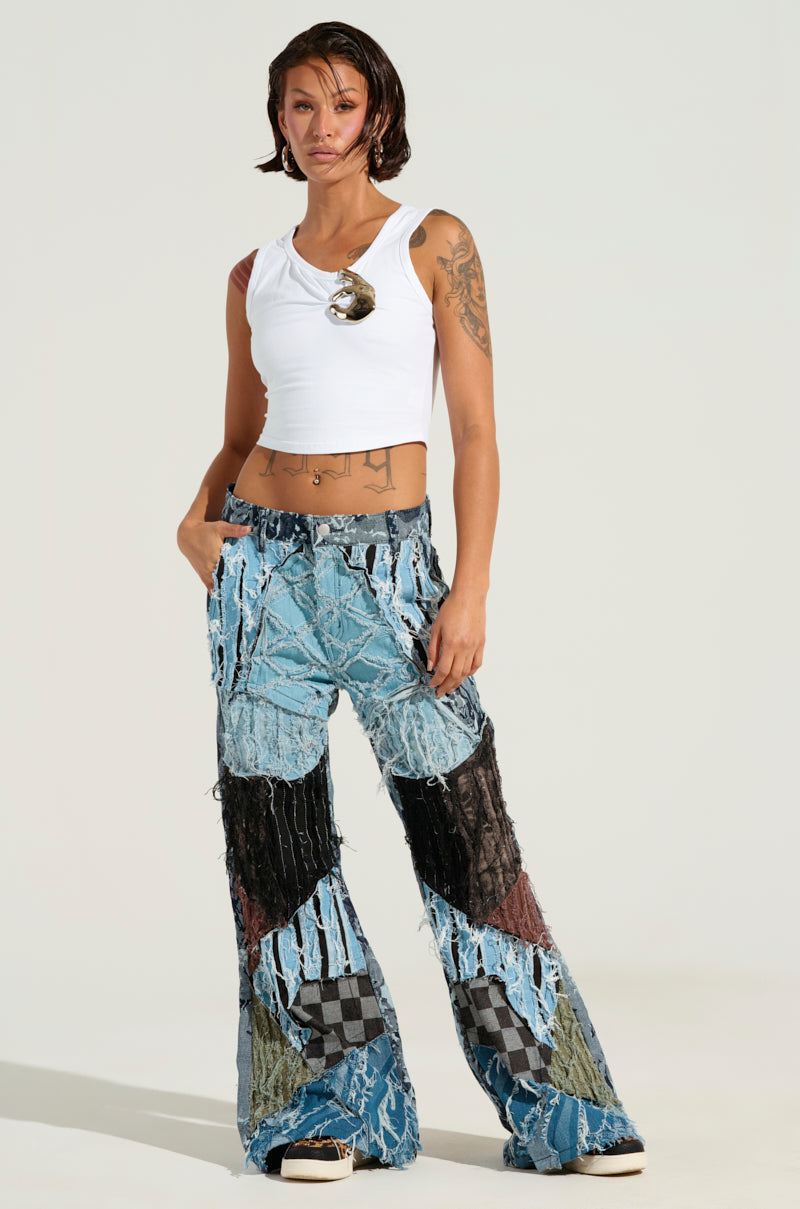A person with short hair stands against a plain background, wearing a white sleeveless top and the RONNY PATCHWORK DENIM jeans featuring a distressed design. Tattoos adorn their arms as they keep their hands in their pockets, exuding a casual and confident style.