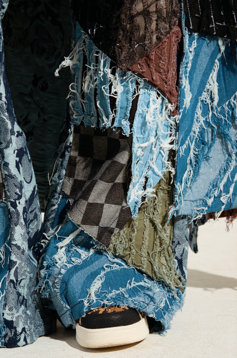 A close-up showcases a person wearing the RONNY PATCHWORK DENIM, characterized by distressed fabric patches such as plaid and lace with frayed edges. Complementing the edgy vibe, a sneaker with leopard print detail is partially visible at the bottom.