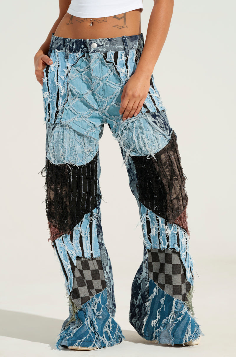 Someone wearing the RONNY PATCHWORK DENIM pants, which showcase an artistic combination of shredded and checkered fabric patterns, stands against a plain background. These distressed jeans feature a blend of various colors and textures, giving them a unique look with a flared leg silhouette.