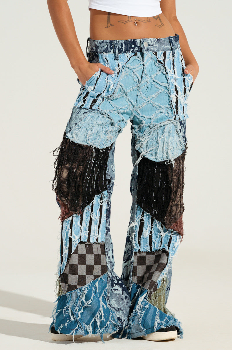 The individual is wearing RONNY PATCHWORK DENIM featuring a distressed design with a mix of denim shades and a checkered pattern. These wide-leg pants are complemented by a cropped white shirt, revealing a tattoo on the waist.