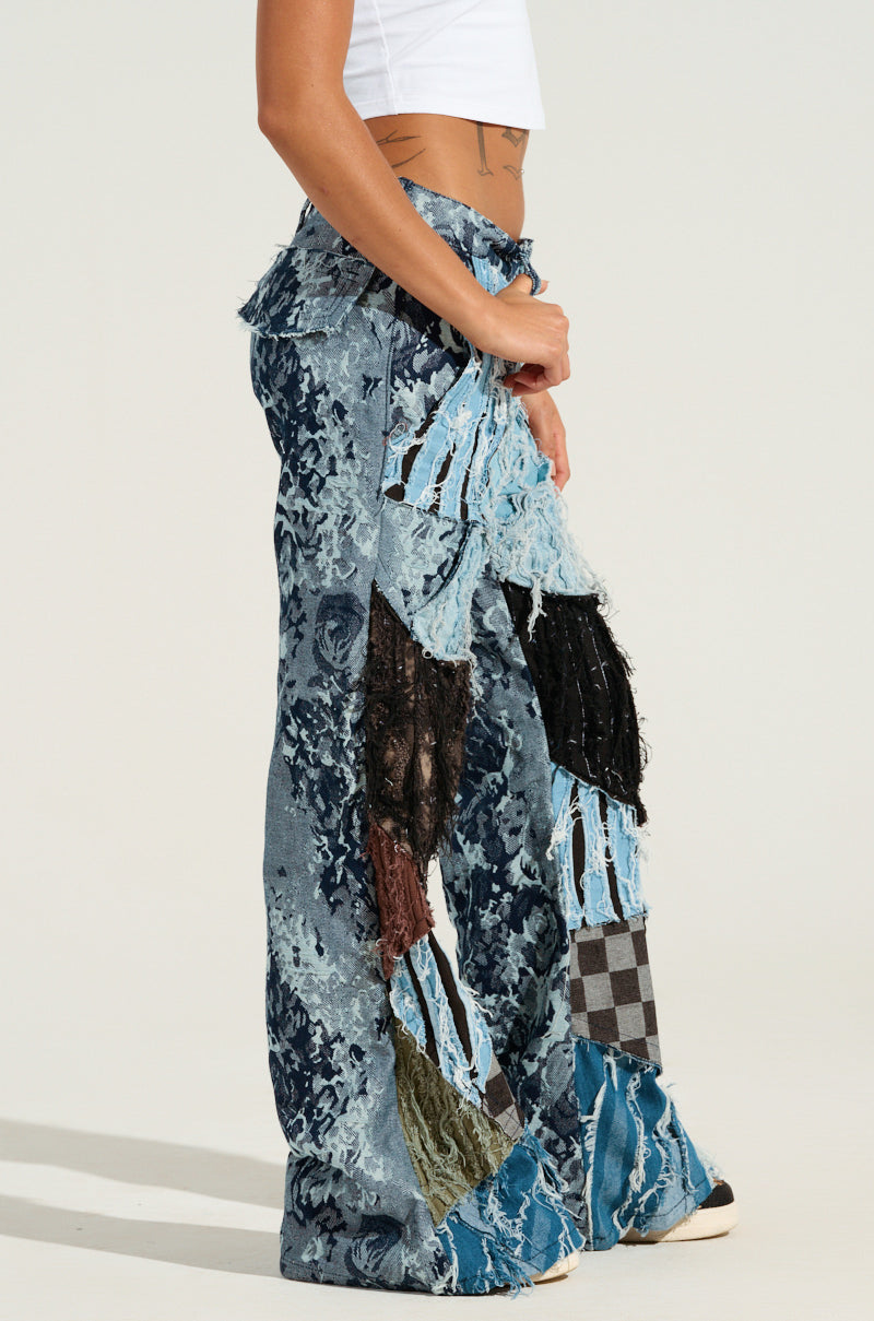 A person wearing the RONNY PATCHWORK DENIM jeans, showcasing an array of denim shades, textures, and patterns, paired with a white crop top. The distressed design highlights frayed edges, lace details, and checkered fabric accents against a plain, light background.