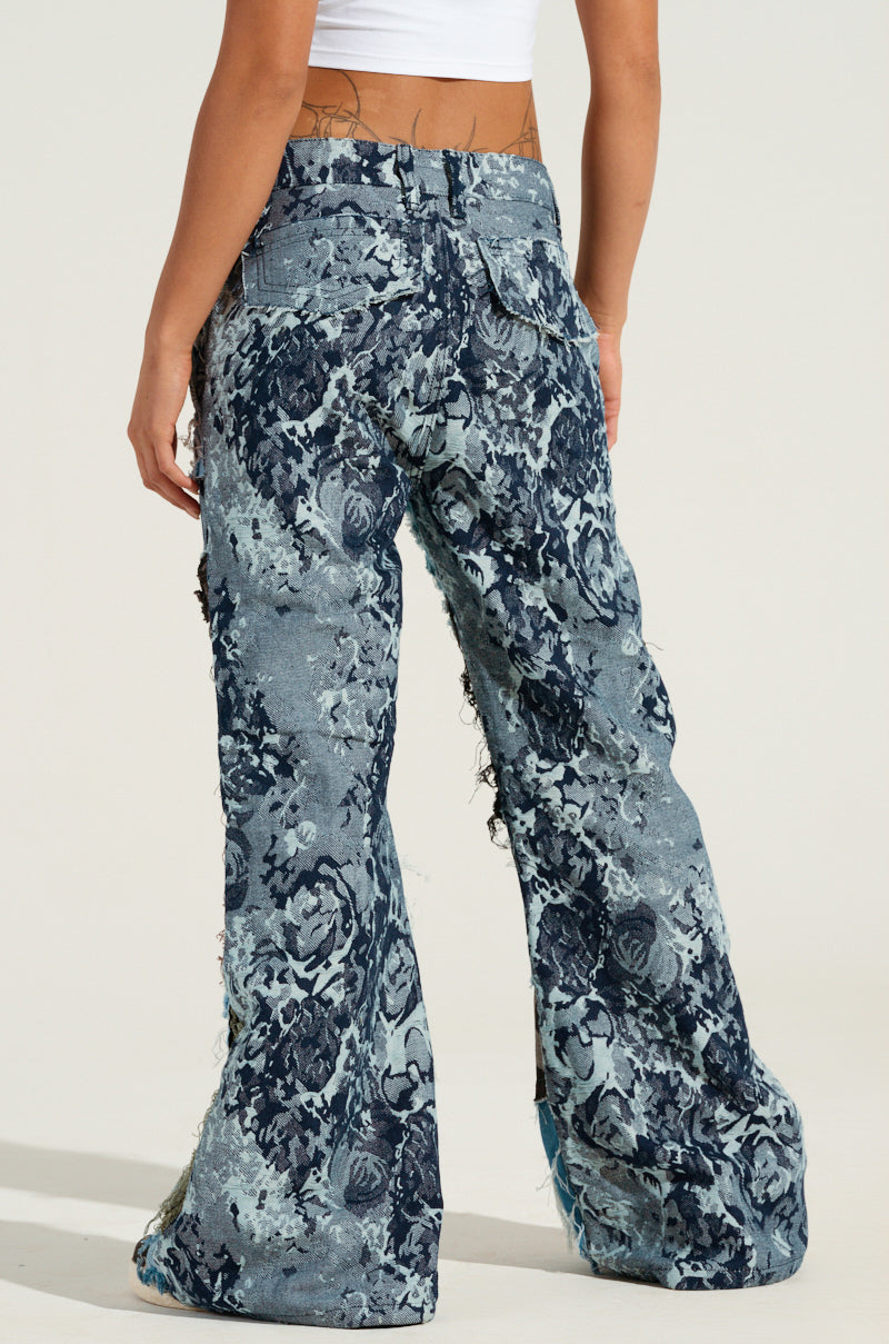 A person in a white crop top showcases the RONNY PATCHWORK DENIM, which features high-waisted, bell-bottomed jeans adorned with a blue and gray floral pattern. These jeans boast a flared leg silhouette with a frayed hem and distressed design, creating a trendy look that highlights their unique style from the back.