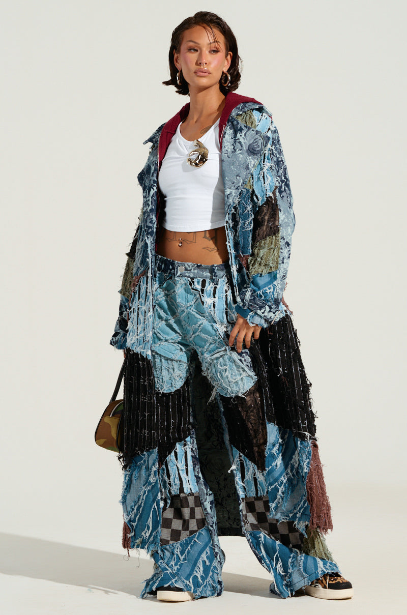 Dressed in the RONNY PATCHWORK DENIM coat and pants, paired with a chic white crop top, an individual highlights a blend of denim shades and patterns with a distressed design. Their look is completed by short hair, statement earrings, and a petite bag against a simple backdrop.
