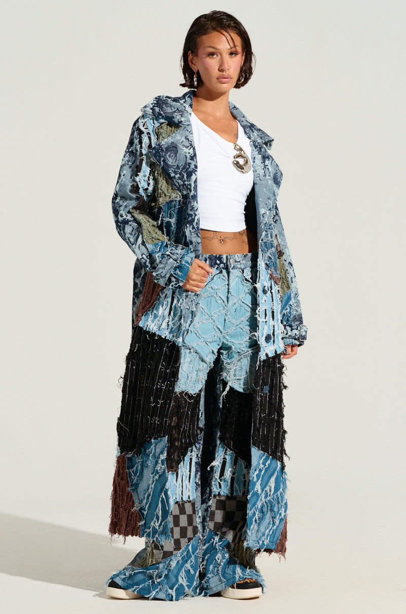 The individual confidently sports the RONNY PATCHWORK DENIM coat and matching pants, both showcasing artful distressed design elements. They pair the ensemble with a white cropped top, sunglasses tucked into their shirt, casual white sneakers, and have short hair.