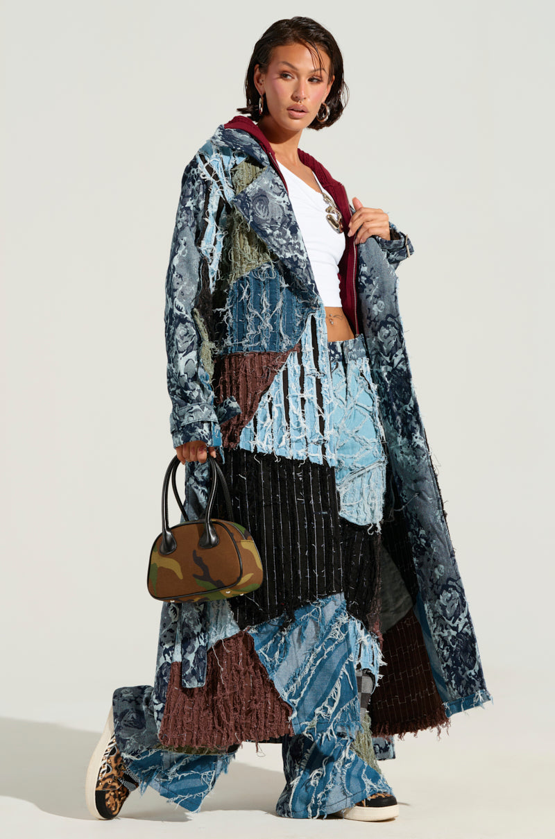 Dressed in the RONNY PATCHWORK TRENCH, a denim coat and matching pants featuring a patchwork design in blue and brown hues are layered over a white top. They carry an accessory - a compact handbag with a camouflage pattern - and finish the look with leopard print shoes. The ensemble is set against a plain, light background.