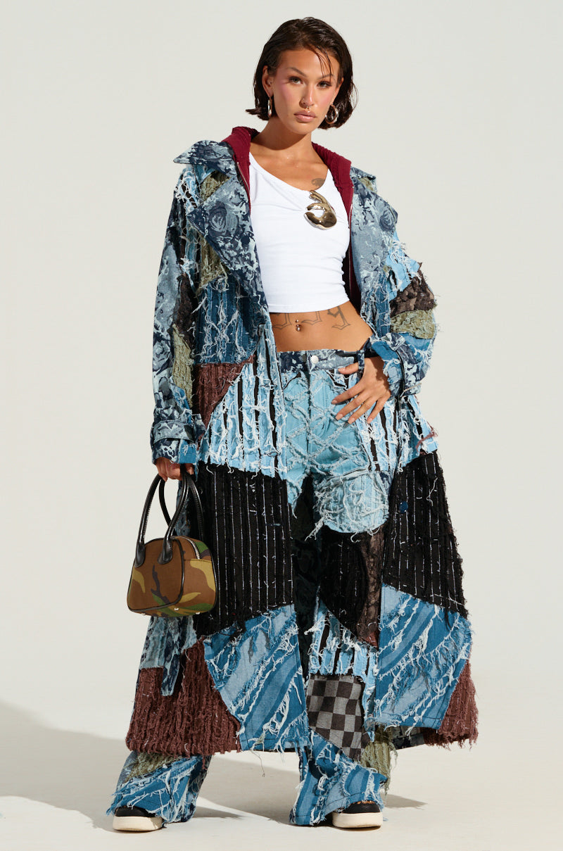 A person stands confidently wearing the vibrant RONNY PATCHWORK TRENCH denim coat and flared pants. They hold a small, camouflage-patterned handbag and accessorize with a large round necklace. The plain background enhances the outfit's intricate details.