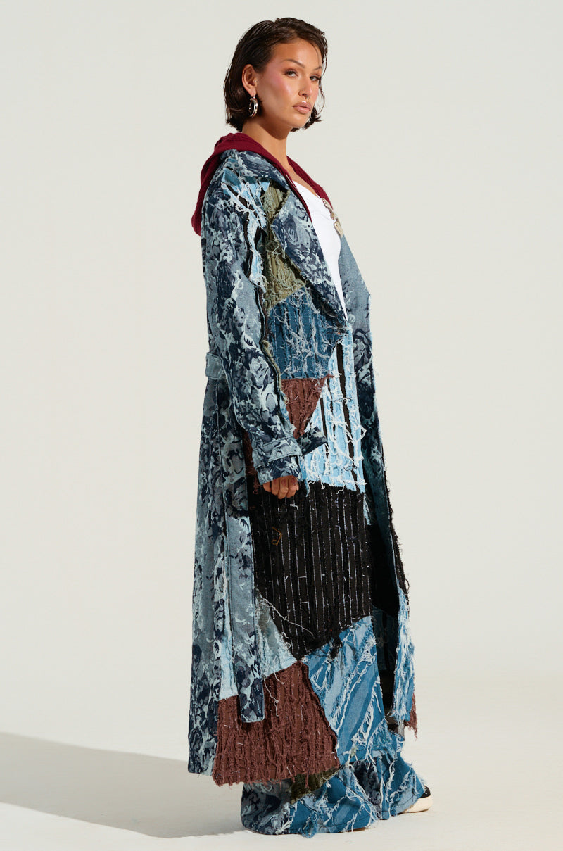 A person is seen in profile, showcasing the RONNY PATCHWORK TRENCH—a striking denim trench coat featuring a patchwork design with various blue-toned patterns and textures. It is paired with matching pants and includes earth-tone accents. The coat also has a hood embellished with subtle floral patterns, set against a plain, light-colored background.