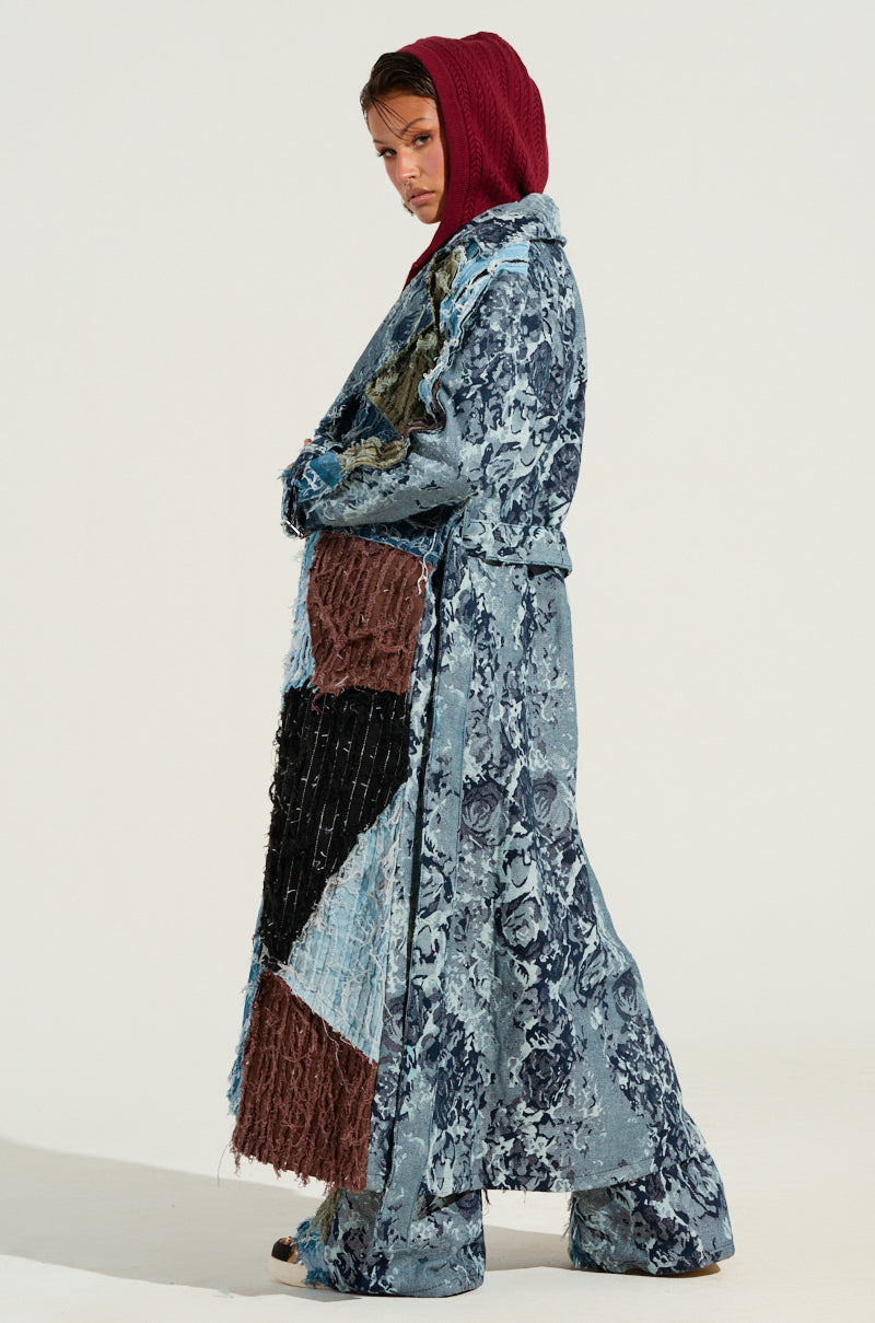 Dressed in the RONNY PATCHWORK TRENCH with its striking blue patterns and large, textured patches, a person in a red hood poses against a plain backdrop, exuding an exquisite, fashion-forward style.