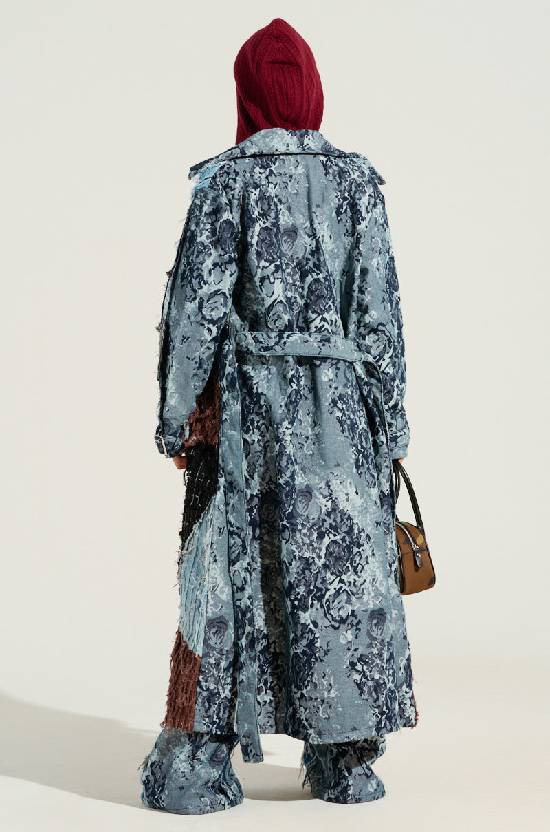 A person is turned away from the camera, dressed in the RONNY PATCHWORK TRENCH with a floral pattern in blue, paired with matching pants and a red knit hat. The ensemble includes boots that seamlessly blend with the pants and is accessorized by a small brown handbag.