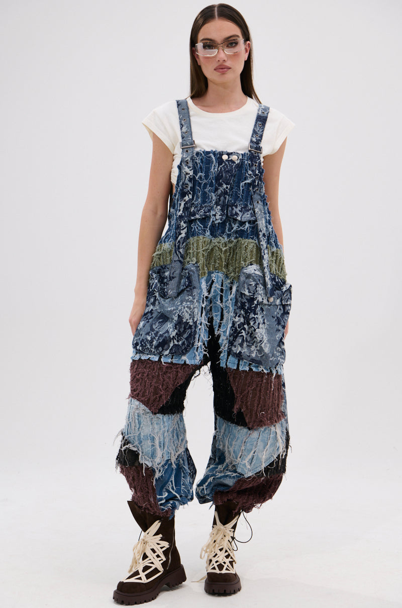 A woman with straight brown hair and round glasses wears the JUST LIKE THAT BAGGY FIT OVERALLS IN PATCHWORK DENIM—featuring blue, green, and brown panels with frayed edges—over a white t-shirt and brown lace-up boots against a plain white background.