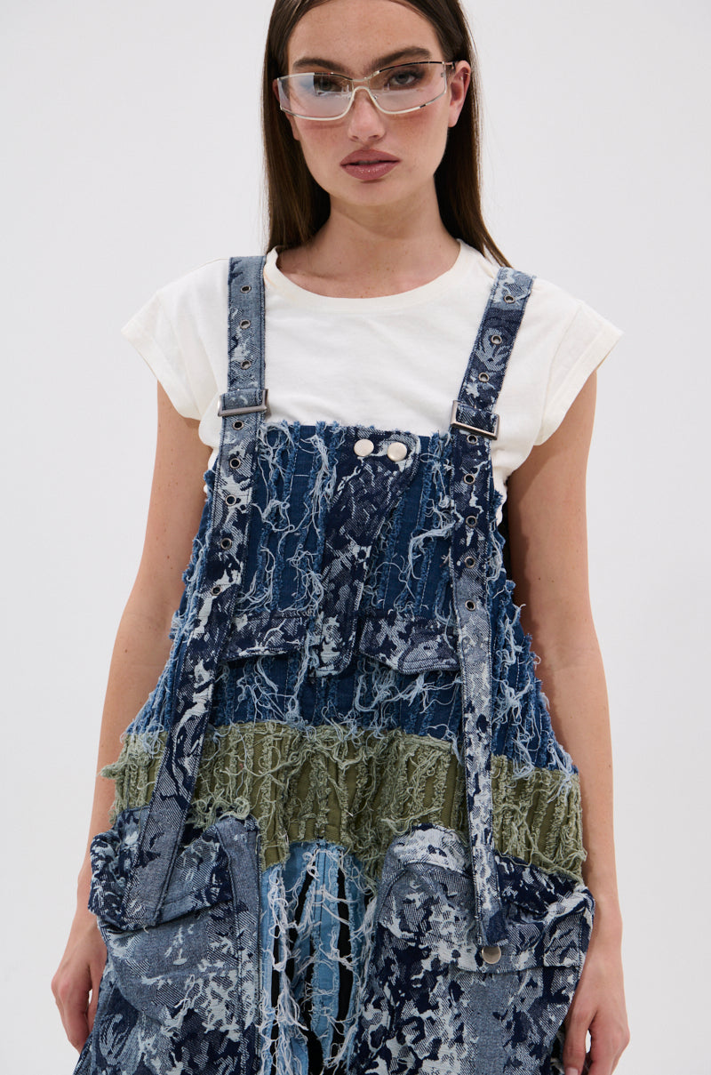 A woman in clear glasses, a white t-shirt, and the JUST LIKE THAT BAGGY FIT OVERALLS IN PATCHWORK DENIM with frayed textures and green accents stands against a plain white background.