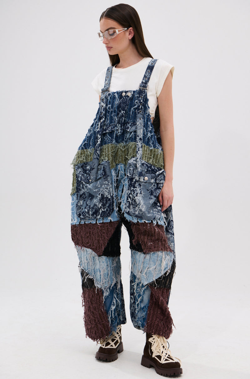 Wearing clear glasses, a white sleeveless top, and the JUST LIKE THAT BAGGY FIT OVERALLS IN PATCHWORK DENIM featuring blue, green, and brown patches, the person stands slightly to the side with chunky beige and brown shoes.