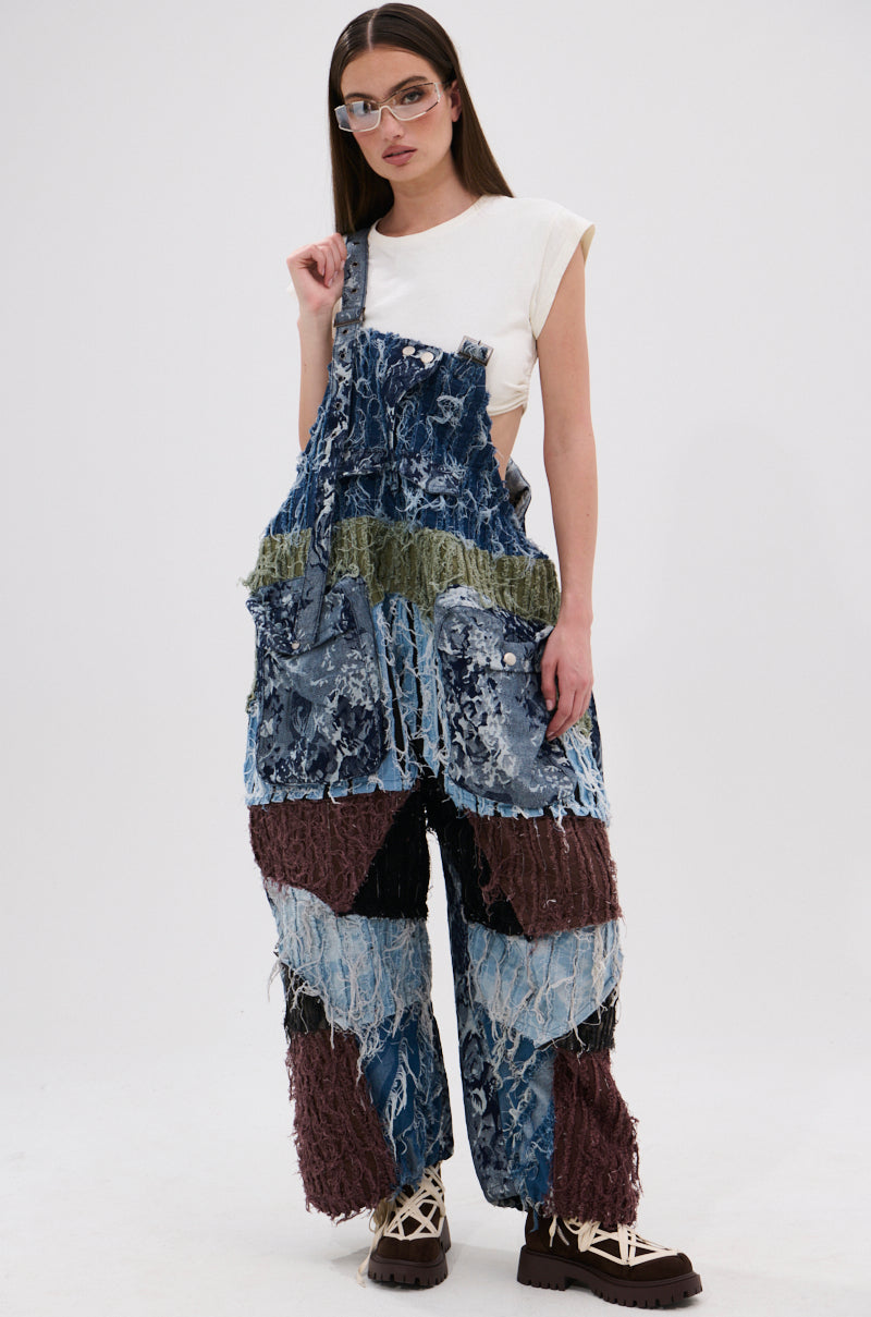 A woman wears glasses, a white sleeveless top, chunky platform shoes, and the JUST LIKE THAT BAGGY FIT OVERALLS IN PATCHWORK DENIM—baggy denim overalls in blue, green, and brown patchwork—standing against a plain white background.
