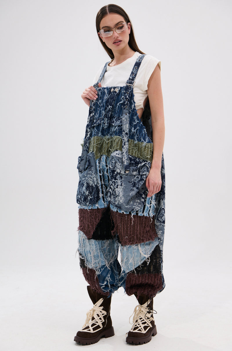 A woman in sunglasses and a white sleeveless top wears JUST LIKE THAT BAGGY FIT OVERALLS IN PATCHWORK DENIM with frayed edges and chunky brown boots, standing against a plain white background.