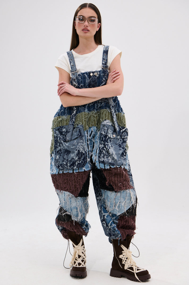 A woman models the JUST LIKE THAT BAGGY FIT OVERALLS IN PATCHWORK DENIM with a white t-shirt, clear glasses, brown lace-up boots, and straight brown hair, posing arms crossed against a plain white background.