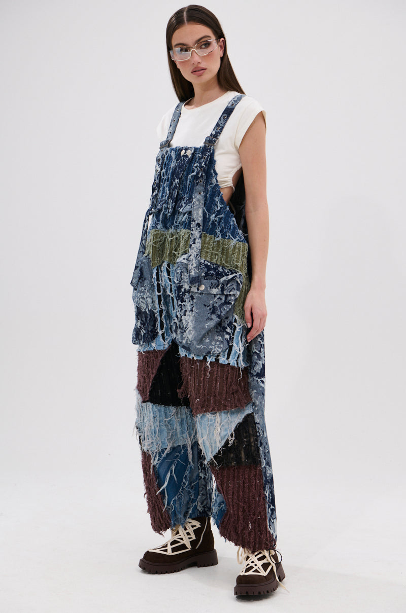 A woman in clear glasses and a white t-shirt models the JUST LIKE THAT BAGGY FIT OVERALLS IN PATCHWORK DENIM, featuring frayed blue, green, brown, and black panels with chunky brown boots against a plain white background.
