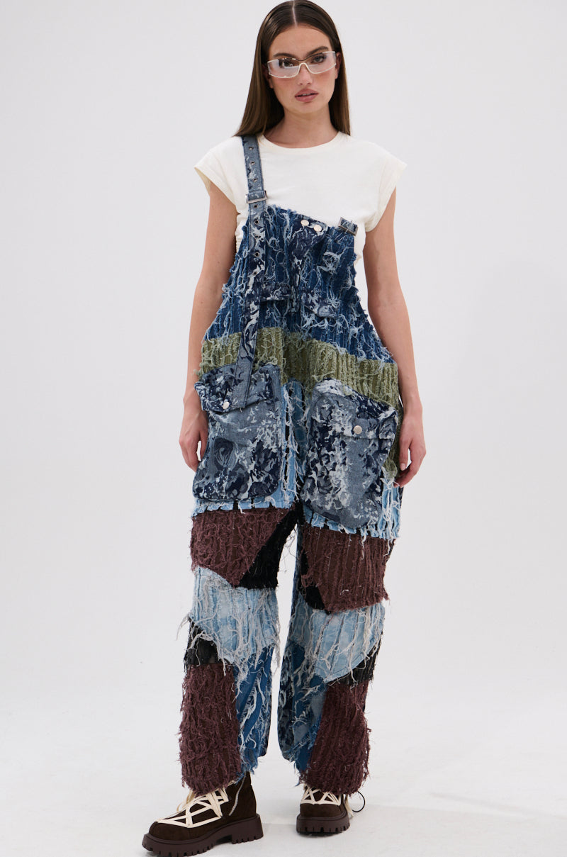 A woman stands facing forward against a white background, wearing sunglasses, a white t-shirt, and the JUST LIKE THAT BAGGY FIT OVERALLS IN PATCHWORK DENIM in blue, green, and brown tones.