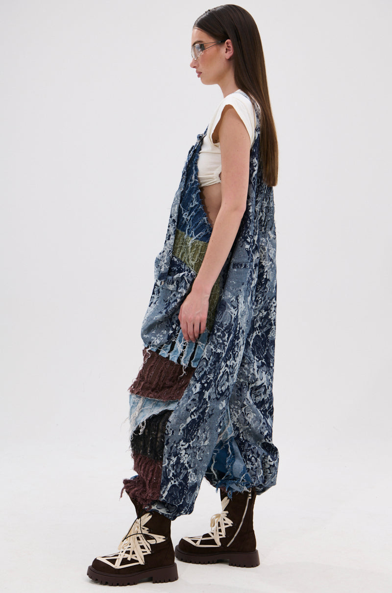 A woman with long brown hair in clear glasses, a white sleeveless top, and chunky brown boots models the JUST LIKE THAT BAGGY FIT OVERALLS IN PATCHWORK DENIM against a plain white background, standing in profile.