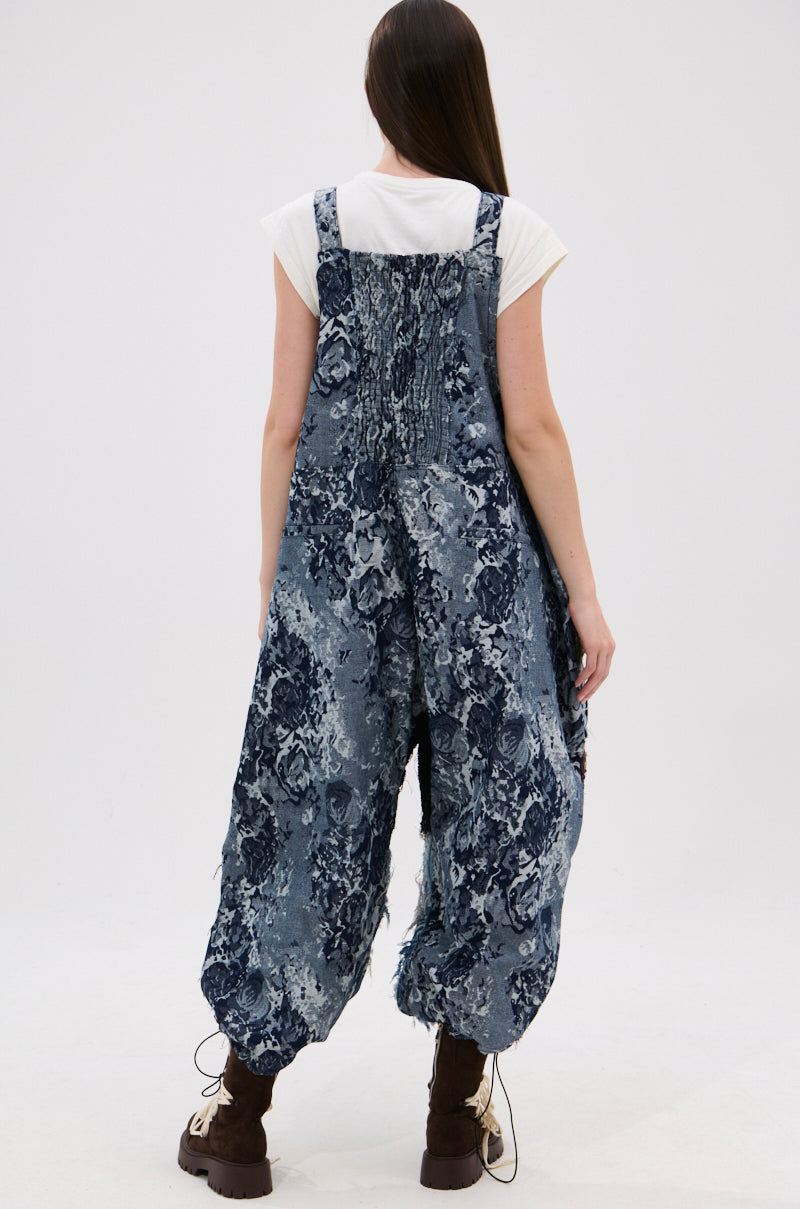 A person with long brown hair, wearing a white short-sleeve shirt and the JUST LIKE THAT BAGGY FIT OVERALLS IN PATCHWORK DENIM, stands facing away from the camera in brown boots against a white background.