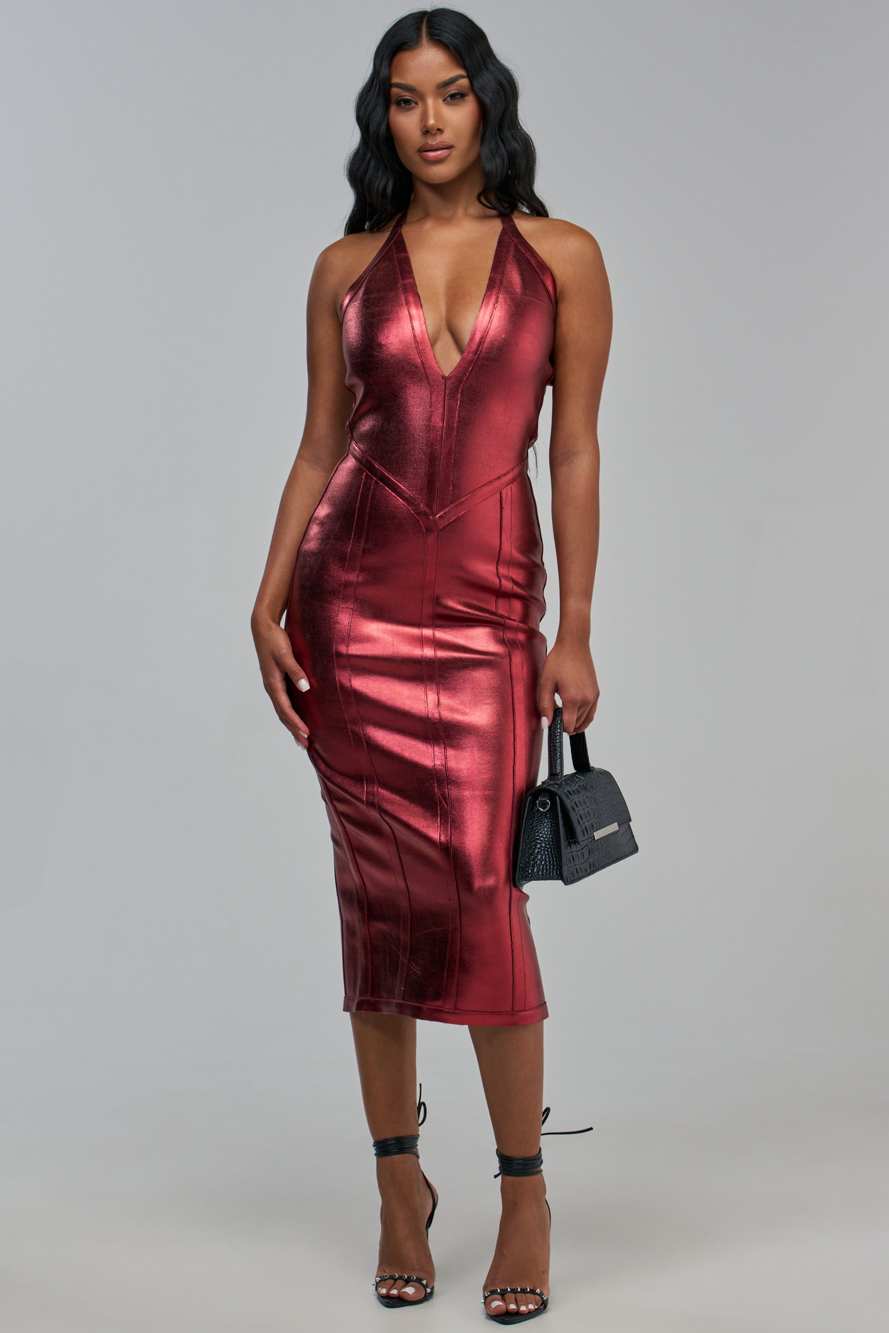 A model wears the TIA BANDAGE MIDI DRESS IN BURGUNDY METALLIC, a burgundy metallic midi dress with a halter neckline and a plunging bodice, paired with black heeled sandals and a small black handbag.