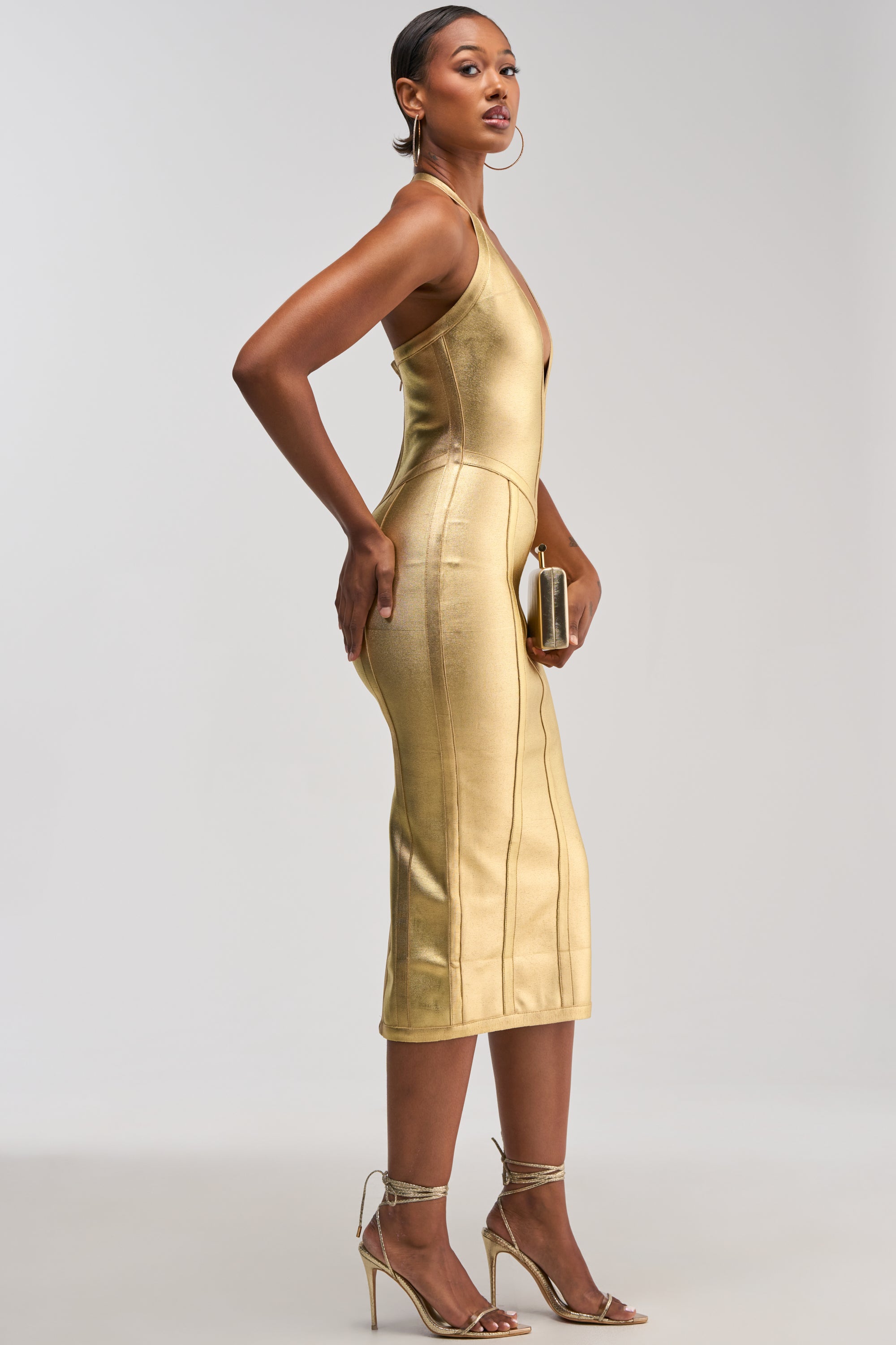 A woman stands sideways against a plain light background, wearing the TIA BANDAGE MIDI DRESS IN GOLD METALLIC with strappy gold heels and holding a small gold clutch.