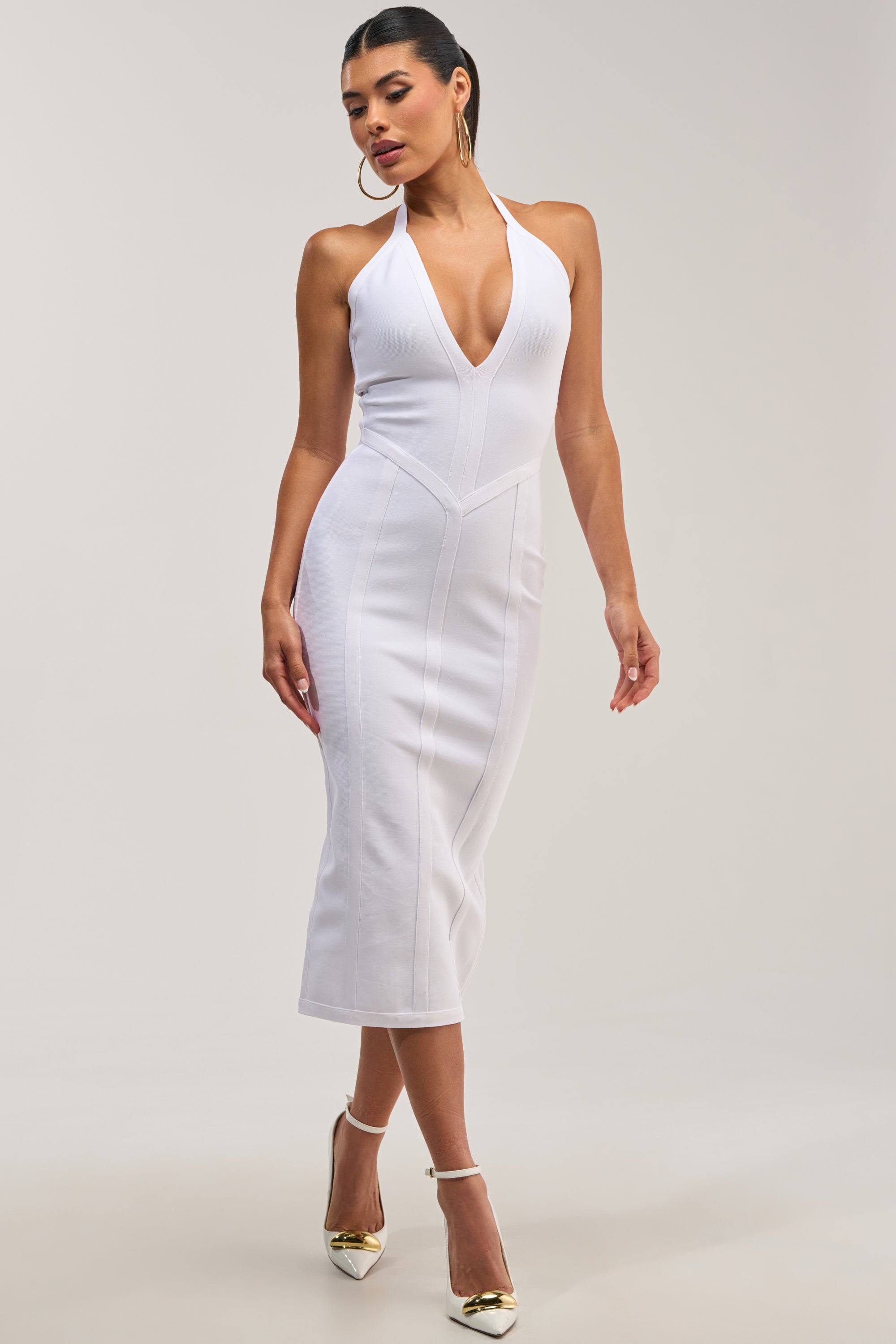 A woman wears the TIA BANDAGE MIDI DRESS IN WHITE, a fitted sleeveless halter piece with a deep V neckline, paired with gold pointed high heels as she poses against a plain background.