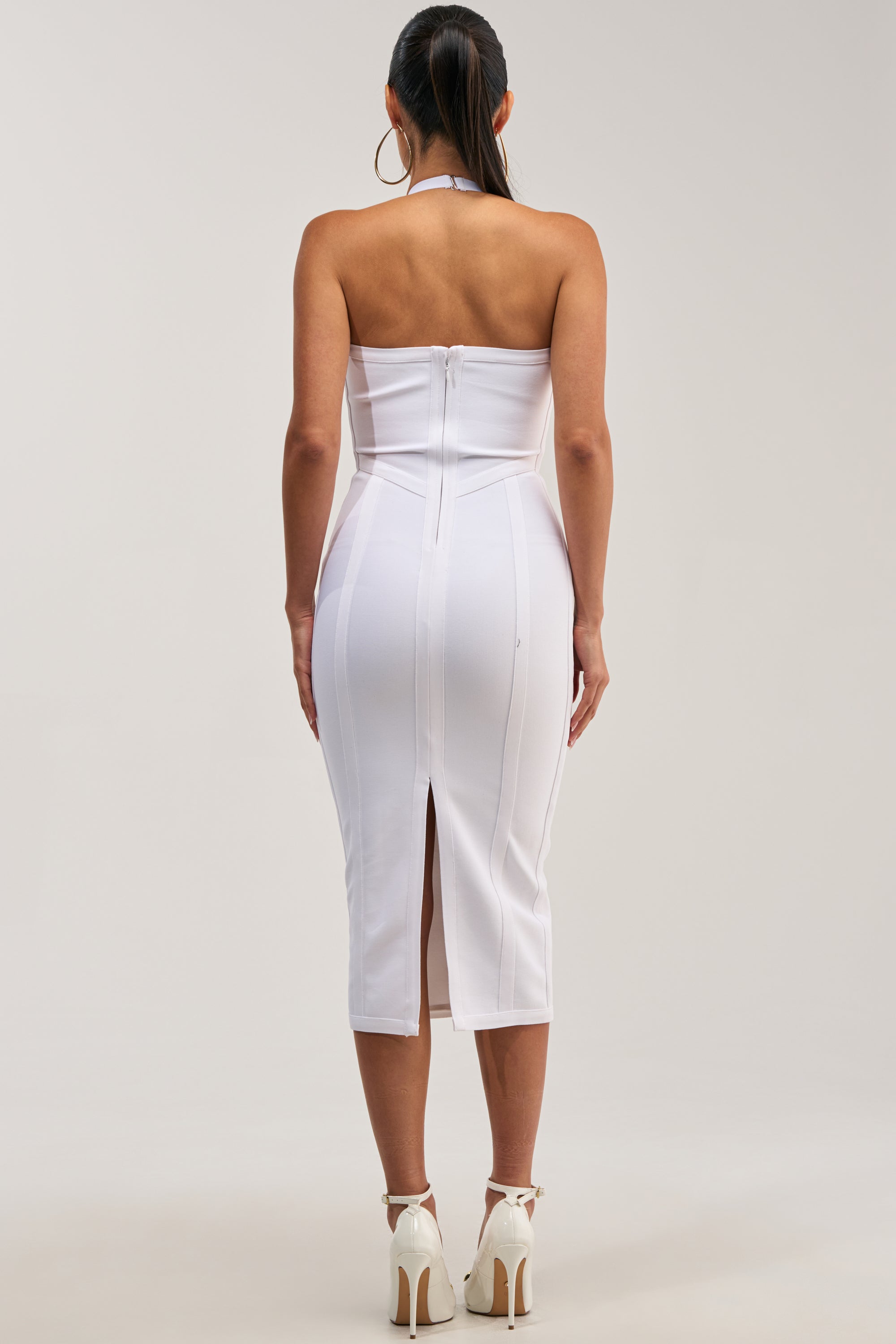 Wearing the TIA BANDAGE MIDI DRESS IN WHITE, a woman stands facing away, showcasing its chic strapless neckline, form-fitting bodycon silhouette, and back zipper with a center slit. She completes her elegant look with hoop earrings, white high heels, and a sleek low ponytail—perfect for any special occasion that calls for making an impression.