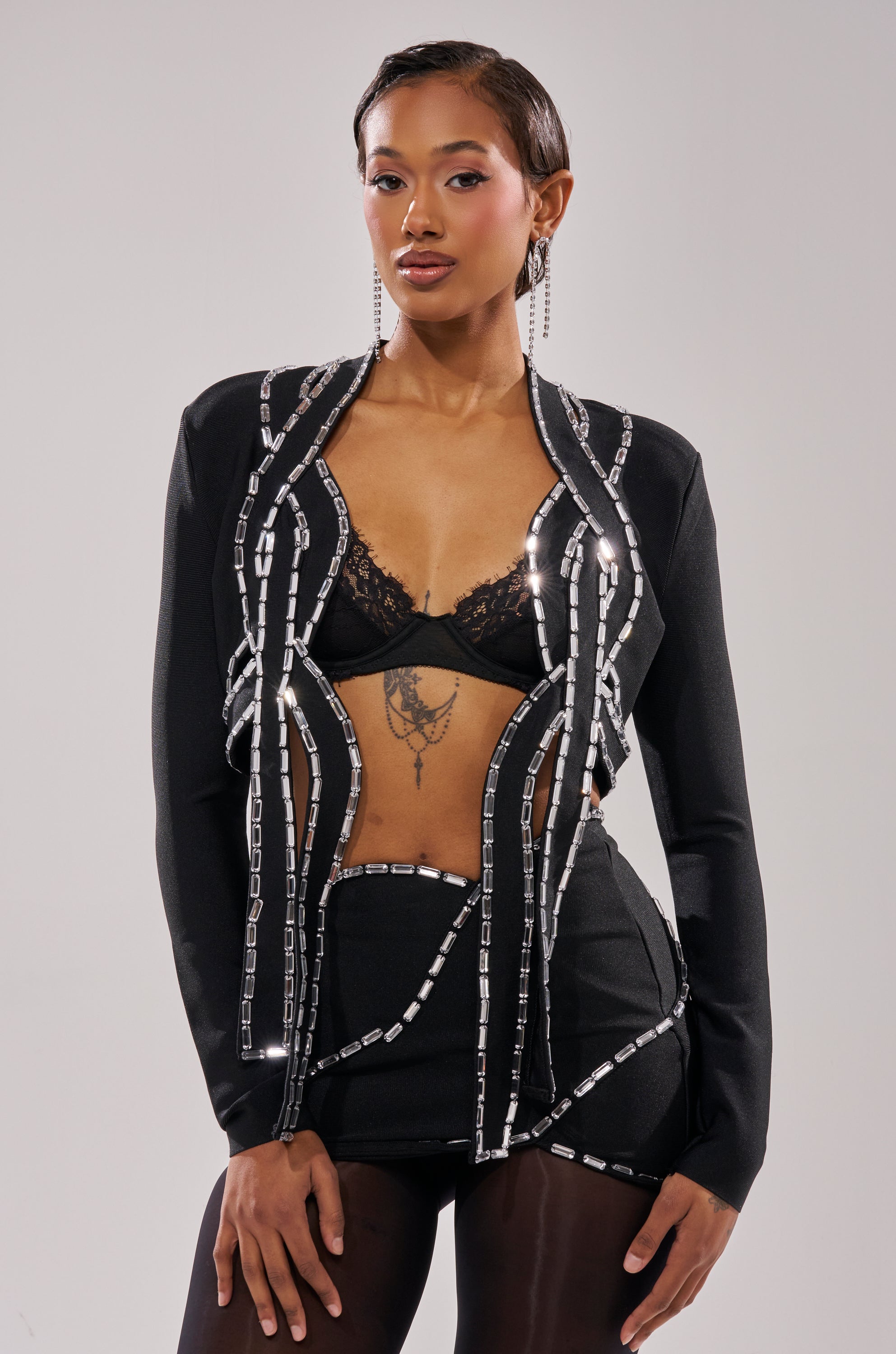 A woman poses confidently in the TOO HOT FOR THIS RHINESTONE BLOUSE, styled with a lacy bralette and high-waisted shorts. She sports short, slicked-back hair, statement earrings, a chest tattoo, and silver chain details on her cropped blouse. 0