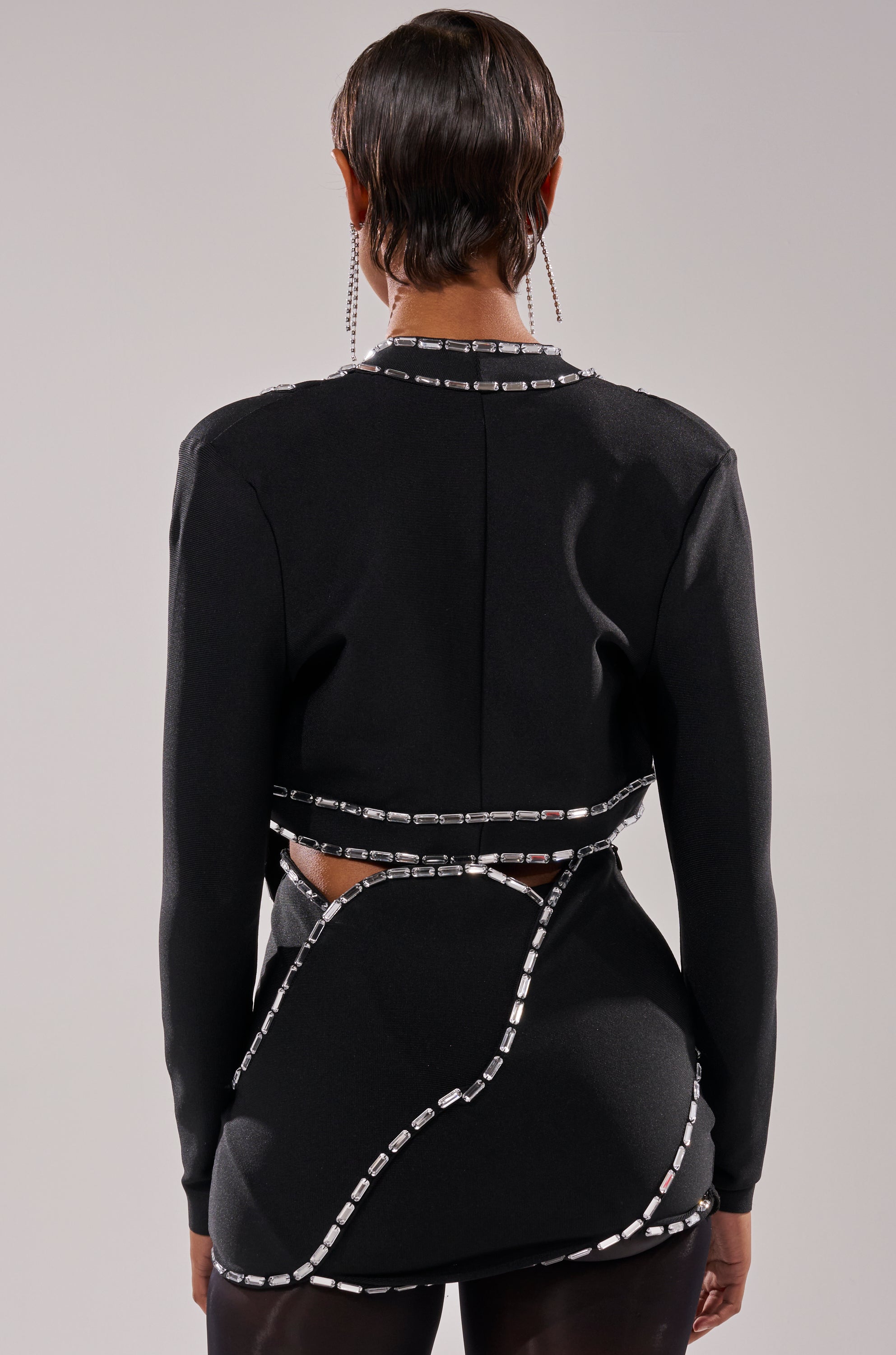 A person with short, slicked-back hair is shown from behind, wearing the TOO HOT FOR THIS RHINESTONE BLOUSE—a fitted black top with silver rhinestone details in geometric patterns. The background is plain and light-colored. 2