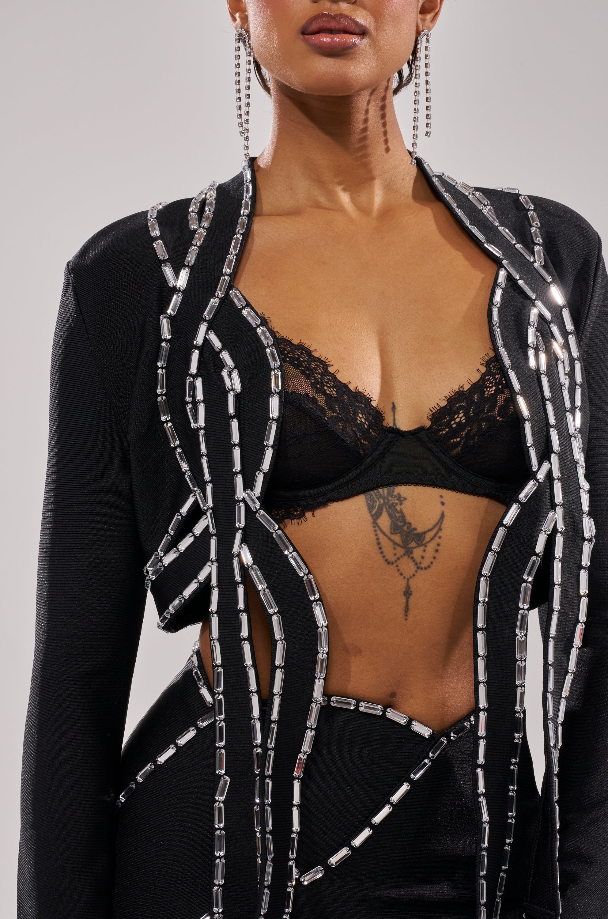 Someone wears the TOO HOT FOR THIS RHINESTONE BLOUSE—black, cropped fit with silver rectangular embellishments—paired with a black lace bralette. Visible are chain and cross tattoos, large hoop earrings, and a partially out-of-frame head. 4