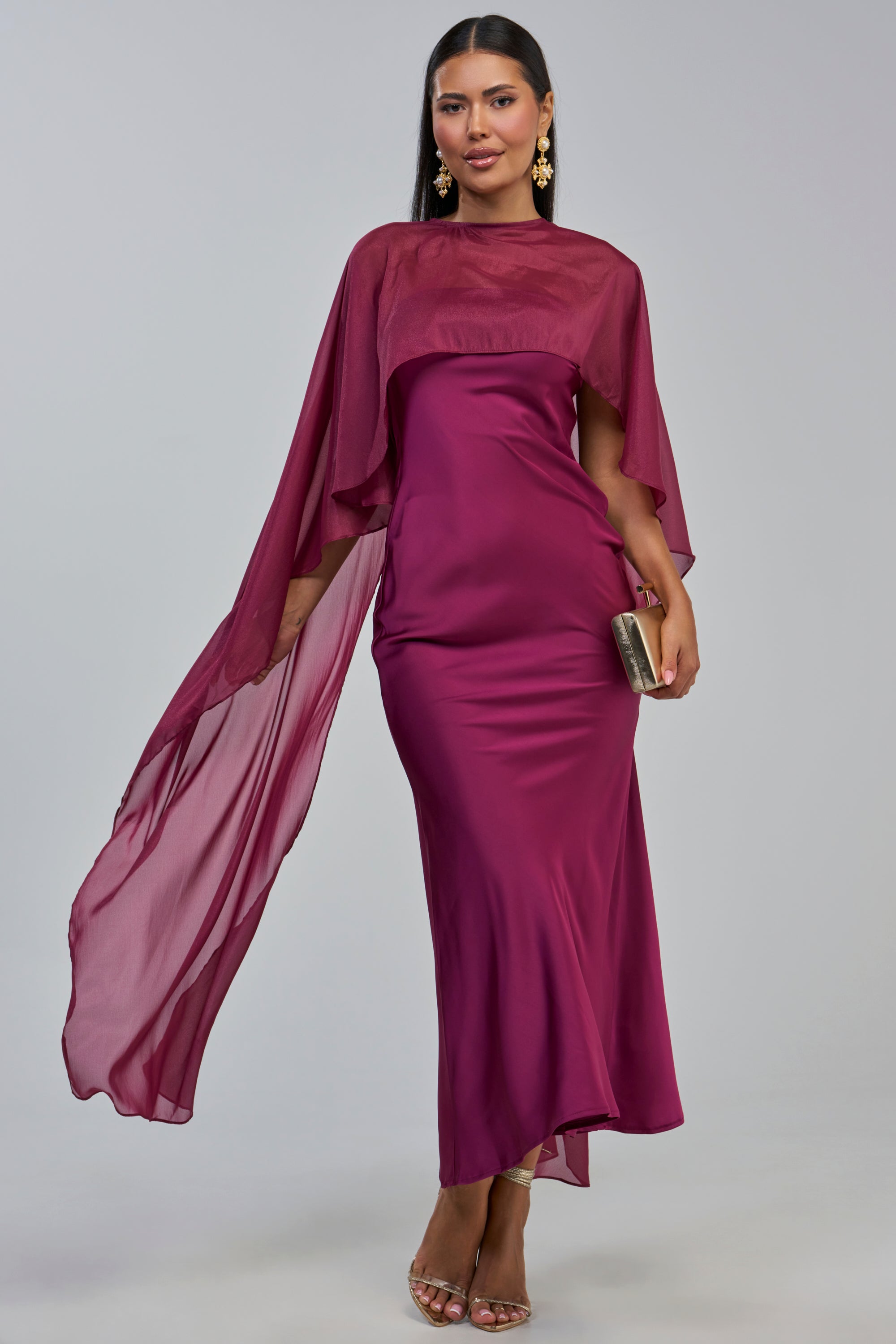 A woman wears the LOUD LUX CAPE SATIN MAXI DRESS—a striking magenta sleeveless, strapless gown with a flowing floor-length silhouette and a sheer cape detail. She accessorizes with gold earrings, a gold clutch, and gold heels as she stands elegantly against a plain light background.