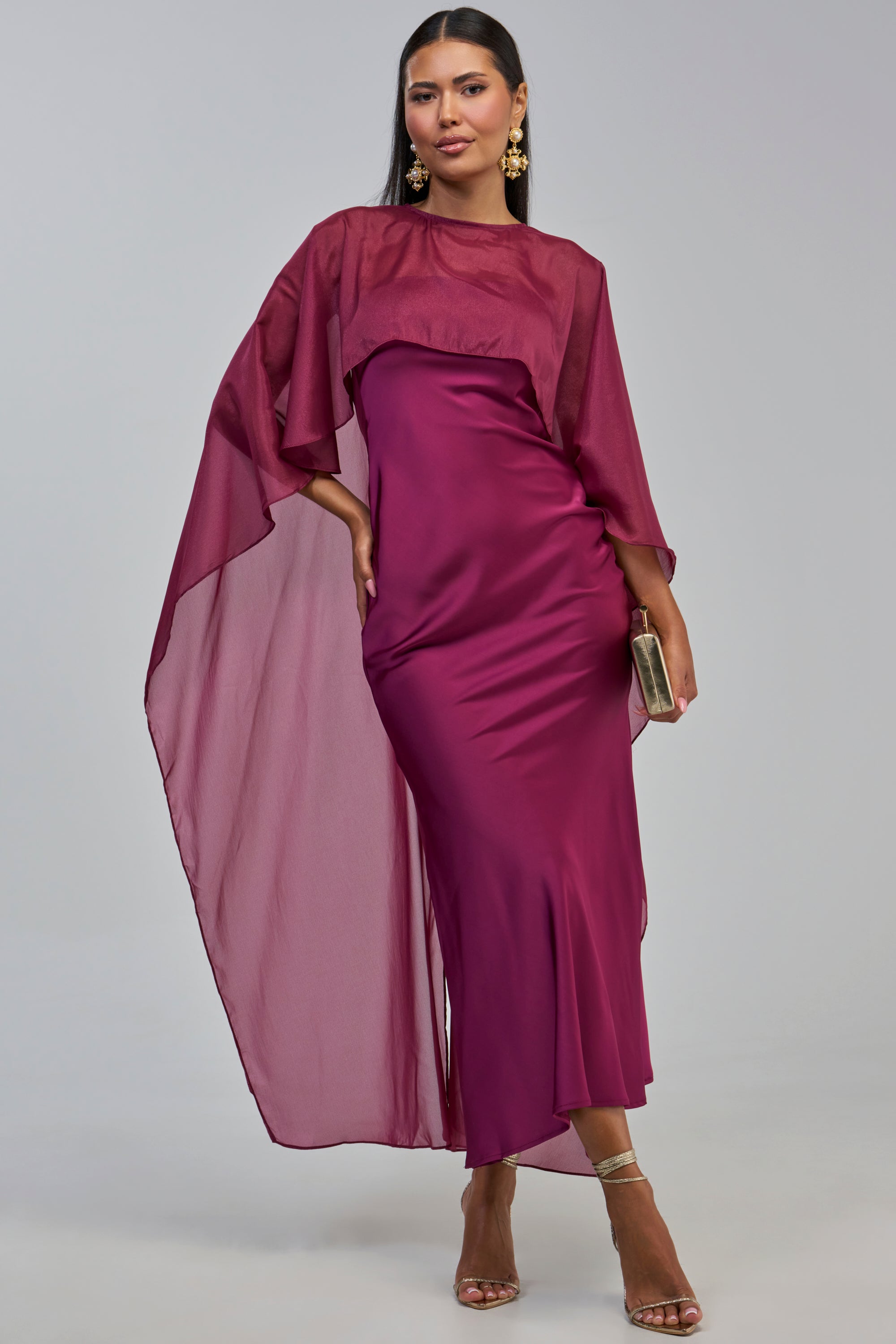 Wearing the LOUD LUX CAPE SATIN MAXI DRESS—a magenta satin gown featuring sheer, flowing cape sleeves—this woman pairs the look with gold heels, statement earrings, and a matching clutch as she stands against a light gray background.