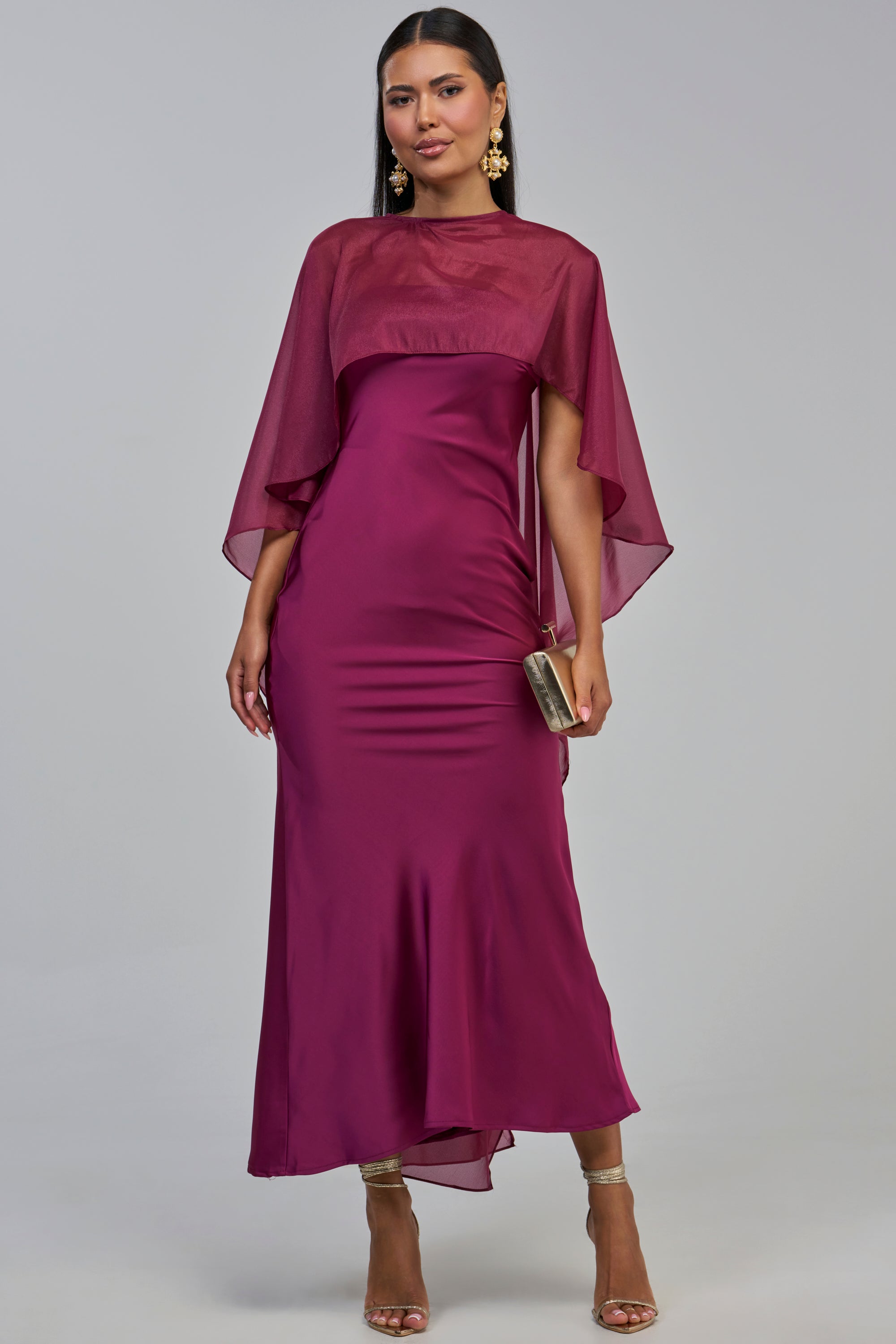 A woman poses against a plain gray background wearing the LOUD LUX CAPE SATIN MAXI DRESS in magenta, which features sheer cape sleeves. She completes her formal evening look with gold earrings, heeled sandals, and carries a small gold clutch.