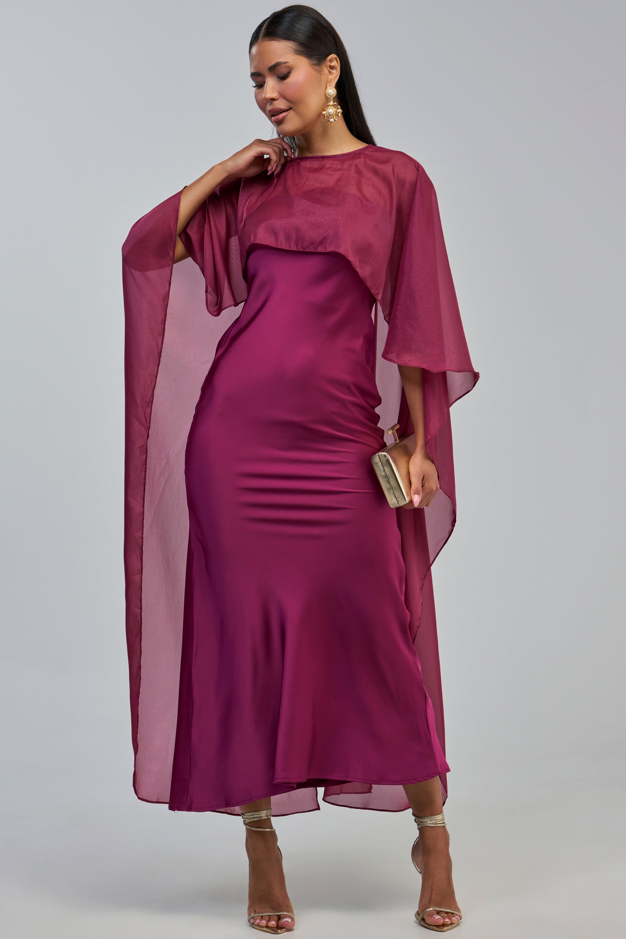 A woman stands against a plain light gray background wearing the LOUD LUX CAPE SATIN MAXI DRESS—a floor-length magenta satin gown featuring sheer cape sleeves. She accessorizes with gold earrings, a gold clutch, and heeled sandals.