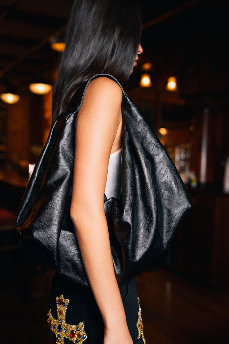 A woman with long dark hair, wearing a white tank top and black embellished pants, carries the EDITORIAL ONLY TOTE over her shoulder in a dimly lit room.