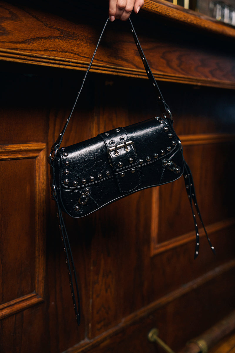 A hand holds the JORDY STUD PURSE, a black bag with silver studs and buckle details in a moto-inspired style, set against a wooden paneled background. 0