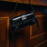 A hand holds the JORDY STUD PURSE, a black bag with silver studs and buckle details in a moto-inspired style, set against a wooden paneled background.