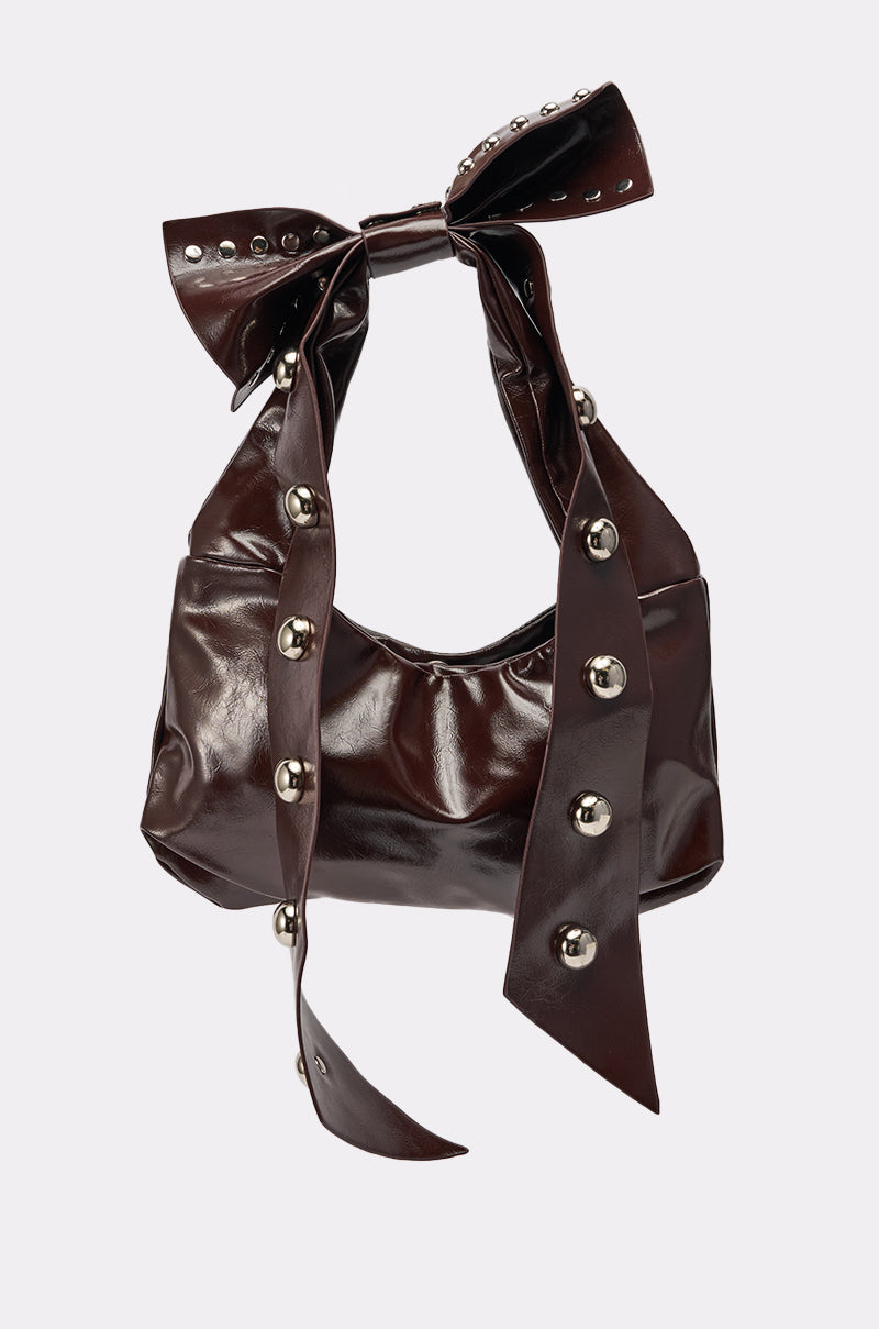 The TIE IT WITH A BOW PURSE is a dark brown, shiny faux leather bag with silver studs and an oversized knotted bow handle, plus long decorative straps with large round studs—a bold statement piece.
