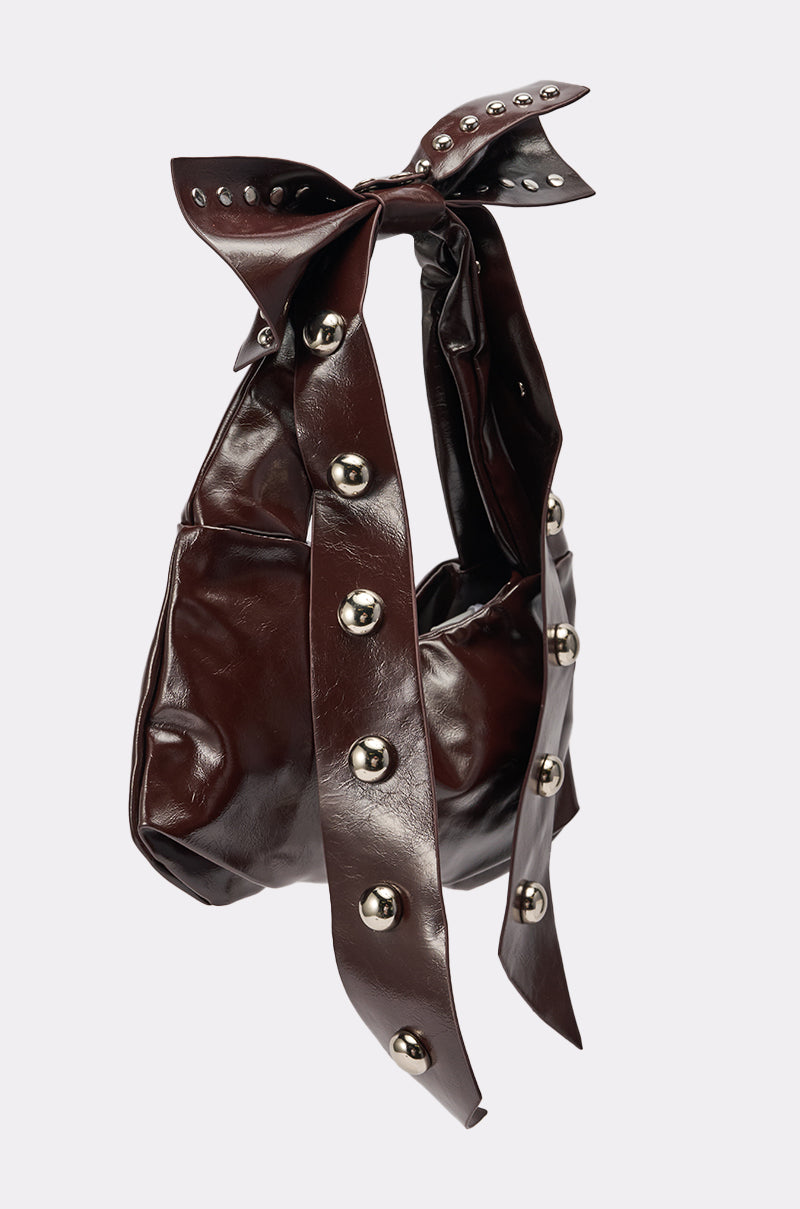 The TIE IT WITH A BOW PURSE is a dark brown oversized handbag featuring a large knotted bow handle and silver studs along the bow and sides, displayed against a plain light background.