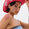 A person with short dark hair sports the ROSES ARE RED COWBOY HAT with white floral designs, a nose ring, and a strapless denim top. A bold maroon stripe tattoo accents their shoulder as they look confidently at the camera.
