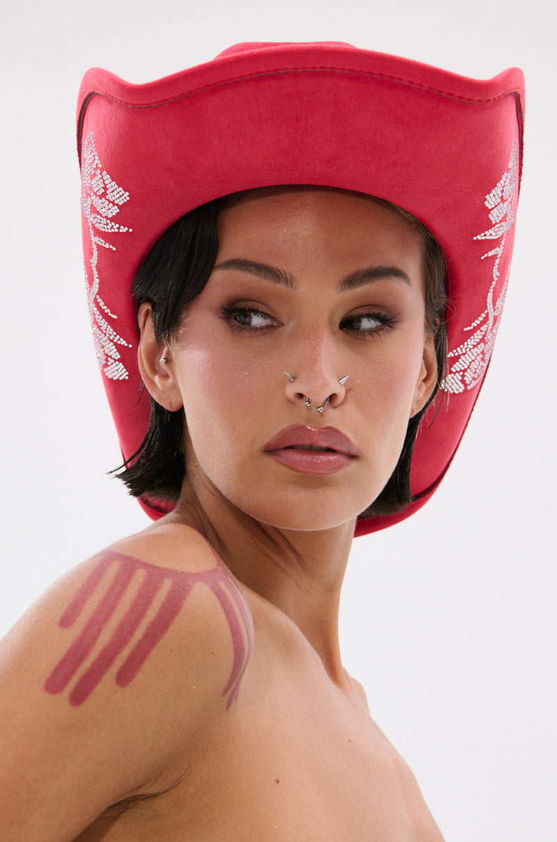 A person with short dark hair and facial piercings wears the ROSES ARE RED COWBOY HAT, featuring rhinestone rose motifs. They display brown makeup swatches on their shoulder against a plain, light background.