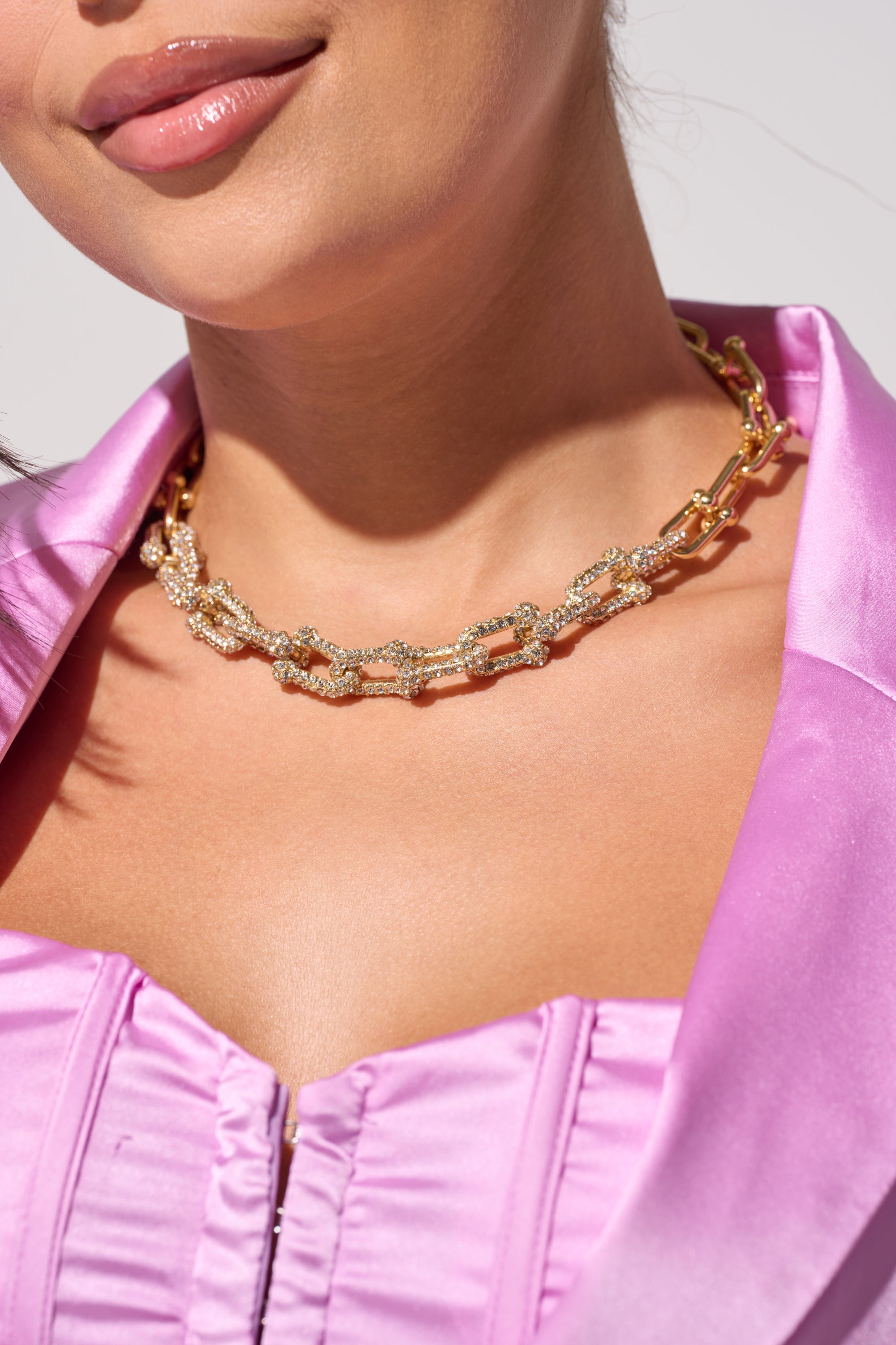 A woman in a satin pink top, partially unzipped, models the LUXE LIVING GOLD NECKLACE with sparkling embellishments against a neutral background. 0