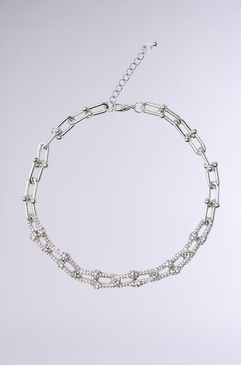 The TIFF NECKLACE features large silver rectangular chain links, accented with sparkling rhinestones, creating a bold statement piece shown on a light grey background. 1