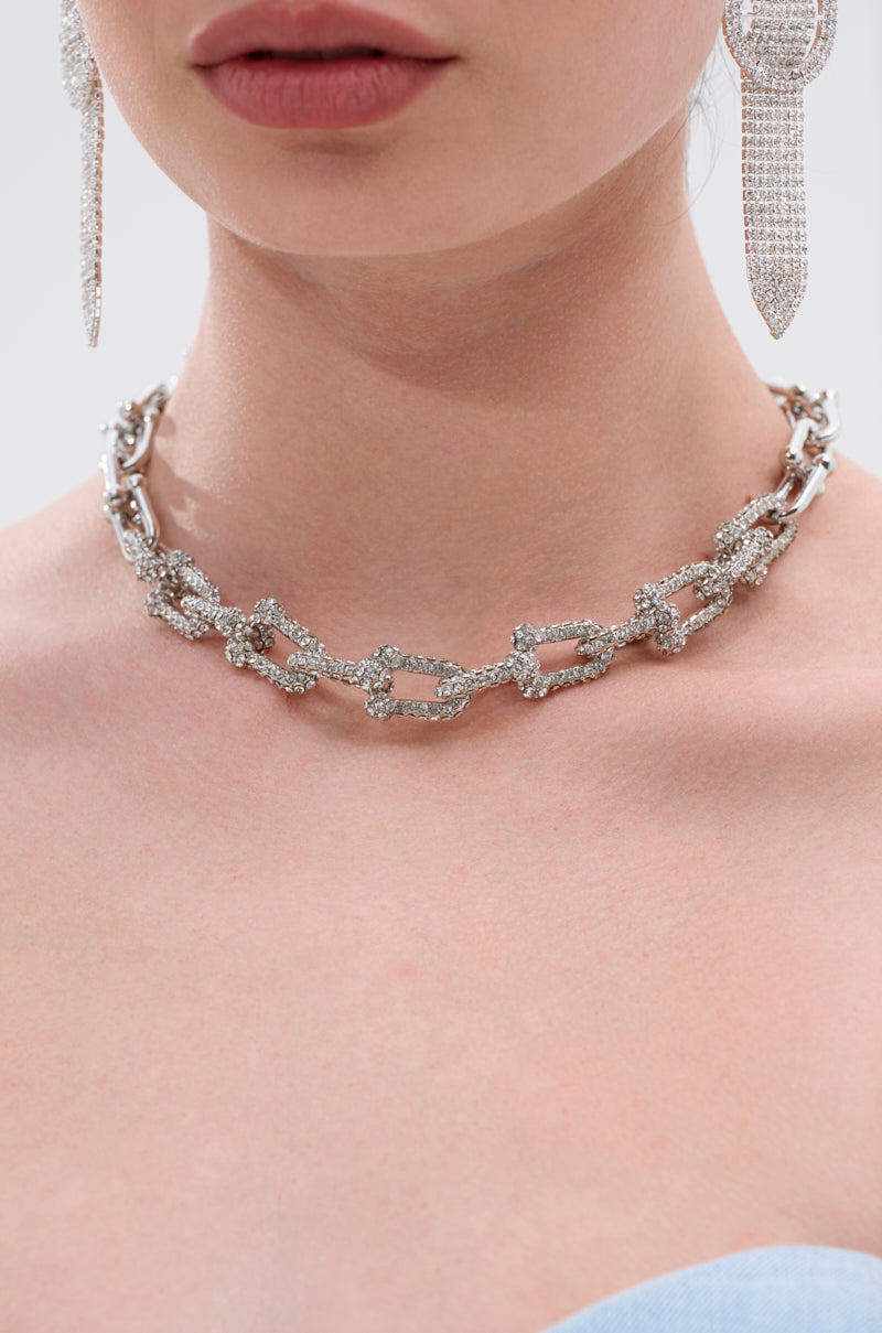 A close-up of a woman wearing the TIFF NECKLACE, a chunky silver chain-link piece, with sparkling dangling earrings. Only her lower face, neck, and shoulders are visible against a neutral background. 3