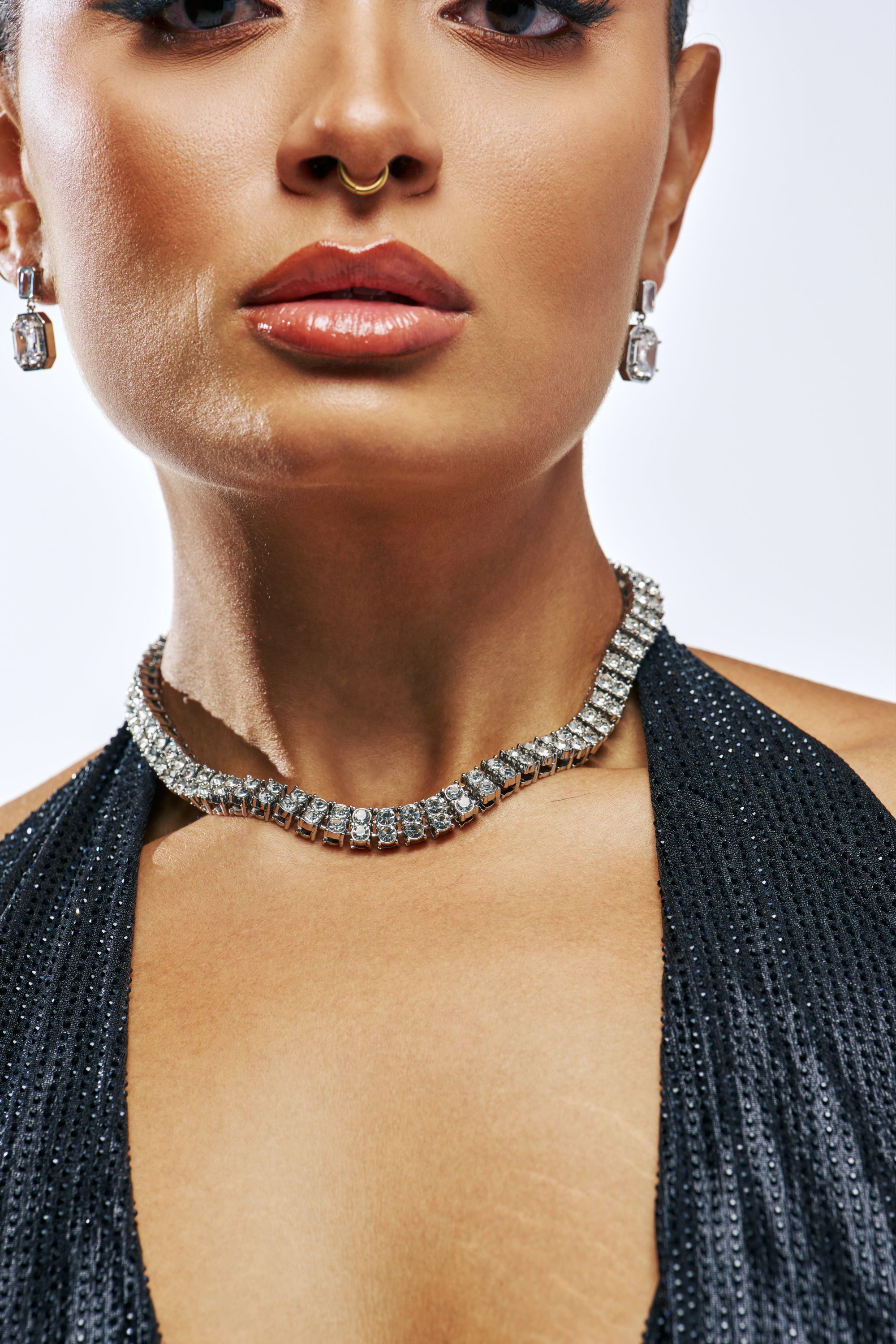 A close-up of a person wearing a sparkling DOUBLE TENNIS NECKLACE, matching drop earrings, and a small gold septum ring.