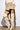 A person dressed in a chic cream long-sleeved coat, cropped white top, and beige ruffled mini skirt showcases the MONTAGE BEIGE STILETTO BOOT against a plain white backdrop.