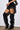 A person wearing a black leather mini skirt with multiple ruffles and the MONTAGE BLACK STILETTO BOOT, featuring a pointed toe, stands against a white background.