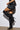 A person wearing a black leather ruffled skirt and MONTAGE BLACK STILETTO BOOTS stands against a white wall. The image highlights the thigh-high boots with an adjustable shaft opening for a perfect fit, focusing on the legs and lower outfit.