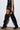 A person confidently strides on a light surface wearing a black outfit with a layered mini skirt and sleek MONTAGE BLACK STILETTO BOOT. The image focuses on their legs, highlighting the pointed toe boots and the stylish, edgy fashion ensemble.
