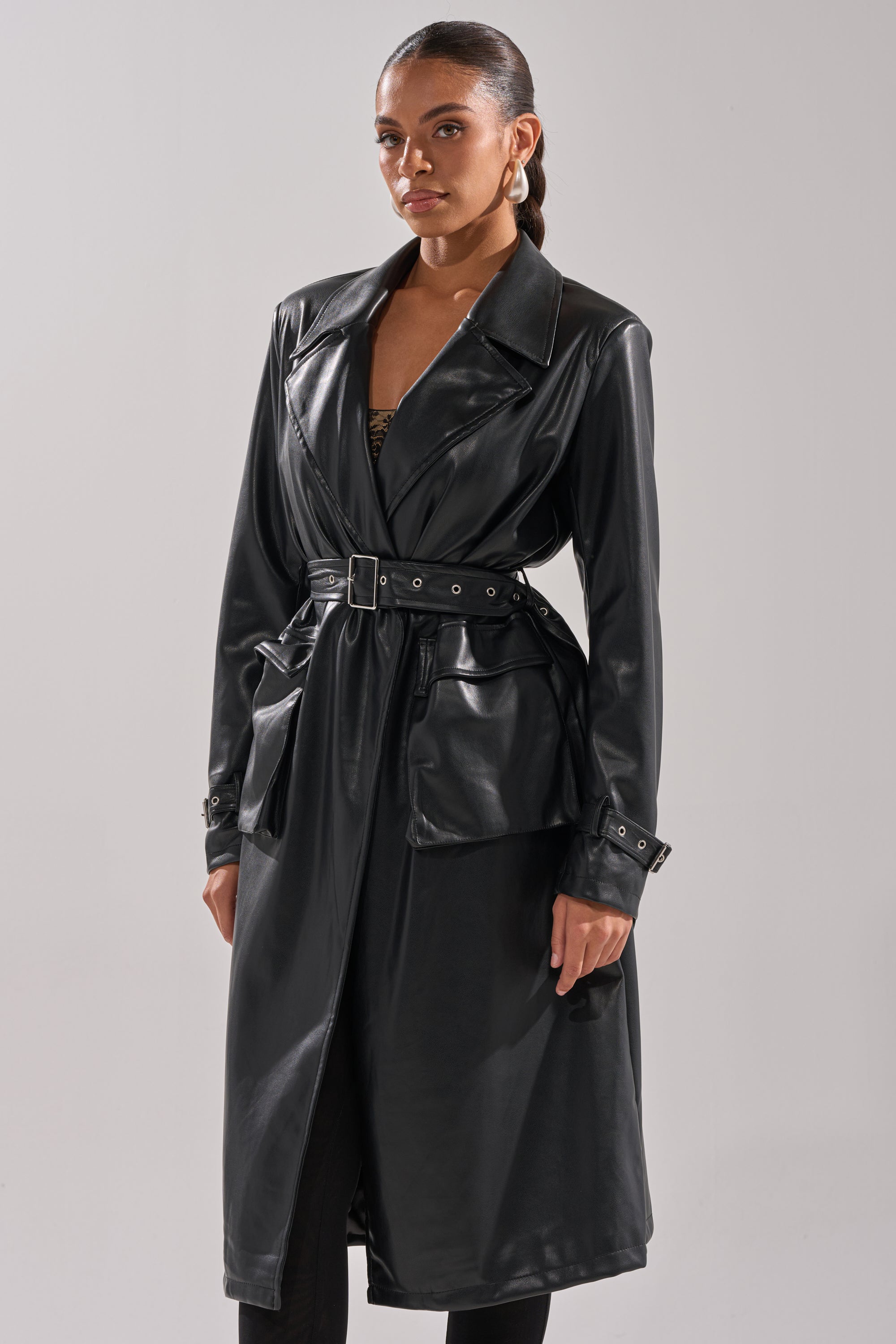 A woman faces forward, wearing the MONTANNA BUTTER LUXE EVERYDAY TRENCH—a long black vegan leather coat with a belted waist, large front pockets, deep collar, shiny finish, and longline silhouette. Her hair is pulled back and she wears hoop earrings. 1